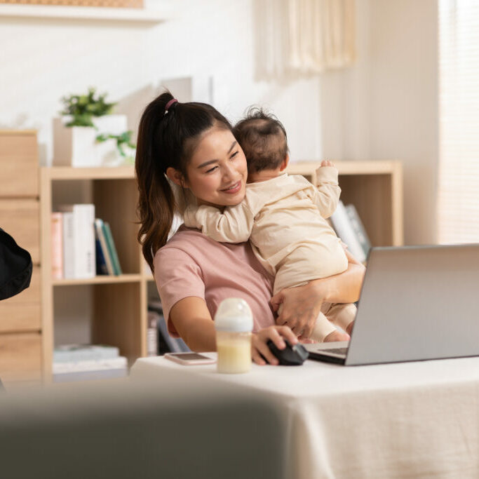 Mother is holding the baby boy while she was working on the laptop. The innocence infant is lean his head on his mom and they both are cuddling. Mom and kid are sitting in the cozy working room.